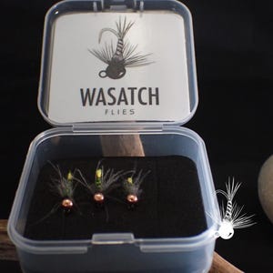May include: A clear plastic box containing three fishing flies. The flies have copper-colored beads, green and yellow accents, and feathery wings. The box lid features the text "WASATCH FLIES" and a fly illustration.