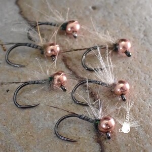 May include: Close-up of six fishing flies. Each fly features a copper-colored bead head, a dark hook, and delicate, feathery wings. The flies are arranged on a textured, light-brown surface, suggesting a natural setting.
