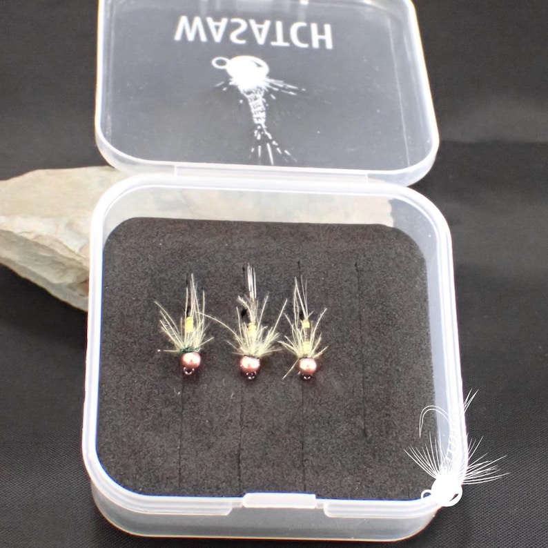 May include: A clear plastic box with the word "WATCH" printed on the lid, containing three fishing flies. The flies have a pink bead head, yellow body, and white feather wings, set against a black foam insert.