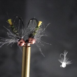 May include: Close-up of two handmade fishing flies. Each fly features a black hook, green body, yellow accents, and silver-gray feathering. The flies are mounted on a gold-colored metal rod against a dark gray background. The flies are designed for fly fishing.