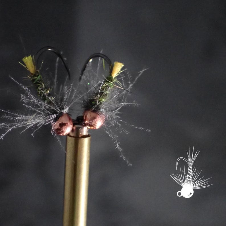 May include: Close-up of two handmade fishing flies. Each fly features a black hook, green body, yellow accents, and silver-gray feathering. The flies are mounted on a gold-colored metal rod against a dark gray background. The flies are designed for fly fishing.