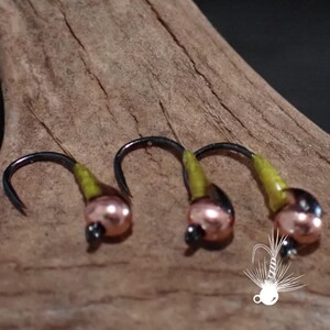 May include: Close-up of three fishing flies. Each fly has a black hook, a copper-colored bead, and a yellow-green body. Fine, light-colored fibers extend from the top of each fly. The flies are arranged on a piece of wood.