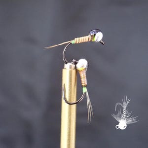 May include: Close-up of two fishing flies. Each fly has a silver bead head, a curved hook, and a body wrapped in tan thread. The flies have small, delicate wings and tails, with green and brown accents. The flies are attached to a gold-colored rod.