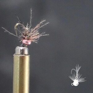 May include: A close-up of a fly fishing lure. The lure has a small, pink bead and a hook with brown and grey feathers. The lure is attached to a brass-coloured metal rod. A white illustration of the lure is in the bottom right corner.