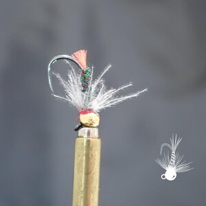 May include: A close-up of a fishing fly with a curved silver hook. The fly features a gold-colored head, white feathers, and a body wrapped in green, red, and white thread. A small orange feather is at the top.