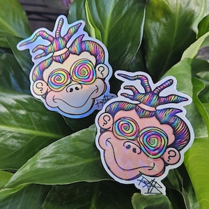 May include: Two die-cut stickers featuring a cartoon monkey face with rainbow-coloured hair styled in a top knot. The monkey has swirling, multi-coloured hypnotic eyes, a small nose, and a smiling mouth. A question mark is visible on the cheek.