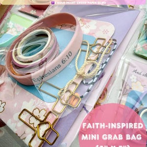 May include: A faith-inspired mini grab bag with 22 items, including a prayer card, notebook, folder, bracelet, stickers, washi tapes, and gold heart cross paper clips. The bag is 4" x 5" and features a pink and floral design. The bracelet has the text "Ephesians 6:10".