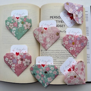 May include: Origami heart-shaped cards with floral patterns in pink, green, and purple, each holding a small card that reads "I love you." The cards are arranged on an open book.