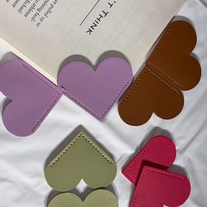 May include: A collection of heart-shaped bookmarks in various colors, including purple, brown, green, and red. The bookmarks are made of a smooth material and are arranged on a white surface. The text "DON'T THINK" is visible on a book in the background.