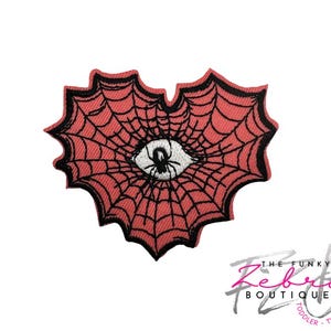 May include: A heart-shaped patch featuring a red spiderweb design with a black outline. The center of the web has a white eye with a black spider. The patch has a black border. The Funky Zebra Boutique logo is in the bottom right corner.