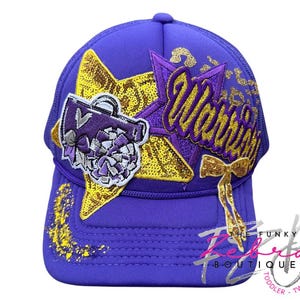 May include: Purple trucker hat with gold sequin star and the word "Warriors" in gold script. Features a cheerleader megaphone patch and a gold bow. The Funky Rebekah Boutique logo is visible on the lower right.
