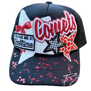 May include: Black trucker hat with a mesh back and a curved brim. The front features the word "Comets" in red, a sequined star, and the phrase "You Had Me At Halftime." Red heart-shaped confetti is scattered on the brim.