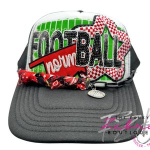 May include: A gray and white trucker hat with the word "FOOTBALL" in black, sequined letters. The hat features a red and white checkered design, a football field graphic, and a red banner that says "norm". A charm hangs from the brim.