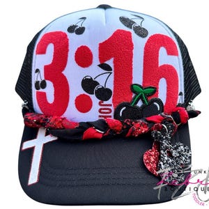 May include: Black and white trucker hat with the number "3:16" in red, adorned with cherry graphics. Features a white cross and a heart-shaped charm. The hat has a braided band with red accents.