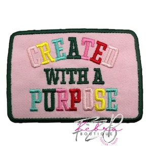 May include: A rectangular pink patch with the words "CREATE WITH A PURPOSE" embroidered in colorful letters. The patch has a green border and a logo in the bottom right corner.