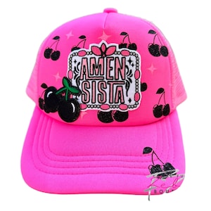 May include: A bright pink trucker hat with a mesh back and a curved brim. The hat features a patch with the words "AMEN SISTA" and a pattern of black cherries. The hat is a vibrant accessory.