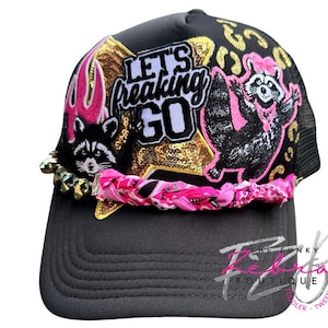 May include: A black trucker hat with a mesh back and a curved brim. The hat features patches of a raccoon, flames, and the text "LET'S FREAKING GO" on a gold sequined star. A pink and black braided band accents the brim.