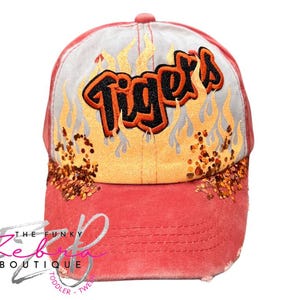 May include: A red baseball cap with the word "Tigers" in orange and black, surrounded by flame graphics. The cap has a distressed look and is embellished with glitter. The Funky Zebra Boutique logo is visible.