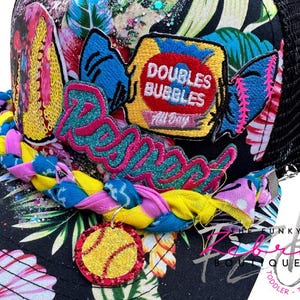 May include: A black trucker hat with a colorful floral pattern and a mesh back. The hat features embroidered patches with the words "Doubles Bubbles" and "Rebel". A softball charm hangs from the brim.
