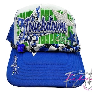 May include: A blue and white trucker hat with the word "Touchdown" in blue script. The hat features a football field design on the crown, embellished with stars and glitter. The brim is solid blue, and a braided band accents the front.