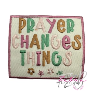 May include: Embroidered patch with the words "PRAYER CHANGES THINGS" in various colors, including green, pink, and gold. The patch has a light background and a pink border, with small flower designs at the bottom. The Funky Zebra Boutique logo is in the bottom right corner.