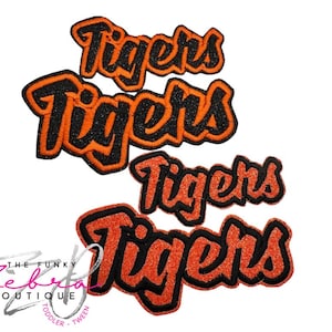 May include: Three orange and black glittery "Tigers" iron-on patches in a script font. The patches have a black outline and are in different sizes. The Funky Zebra Boutique logo is in the lower left corner.