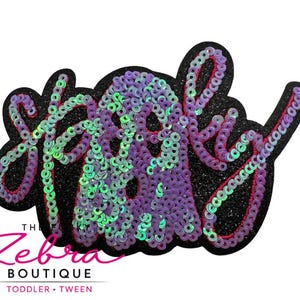 May include: A black glittery patch with the word "Spooky" in a cursive font. The word is filled with purple and green sequins. The patch has a ghost shape in the center. The Zebra Boutique logo is at the bottom.