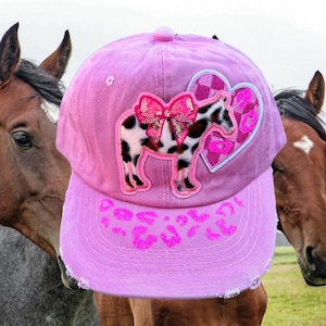 May include: Pink baseball cap with a horse and heart design. The cap features a horse applique with a sequined bow and a heart-shaped patch. The brim has a pink leopard print pattern. The background includes two horses.