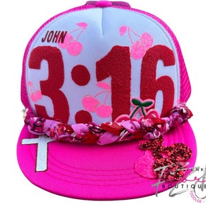 May include: A bright pink trucker hat with a white front panel. The hat features the text "JOHN 3:16" in red, with cherry illustrations. A decorative braided band and a cross are on the brim. A glittery cherry design is on the side.