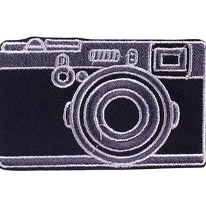 May include: A black and gray embroidered patch in the shape of a vintage camera. The patch features detailed stitching outlining the camera's body, lens, and various buttons. The design is a classic representation of a retro camera.