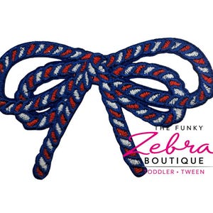 May include: A blue, red, and white embroidered bow. The bow is made of a thick, rope-like material and features a repeating pattern of red and white zigzags against a blue background. The Funky Zebra Boutique logo is visible.
