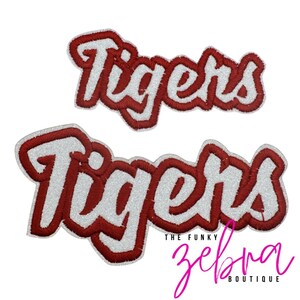 May include: Two embroidered patches featuring the word Tigers in a white glittery script font with a thick dark red border. The patches are arranged with one smaller version positioned above a larger version against a plain white background.