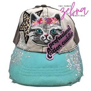 May include: A distressed baseball cap with a cat face patch wearing a floral crown. The cap features a turquoise brim, a leopard print star, and the text "professional kitten cuddler". Embellished with sequins and rhinestones.