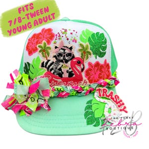 May include: A mint green trucker hat with a white mesh back and a decorative front panel. The panel features a raccoon on a flamingo floatie, tropical flowers, and the word "Summer". The hat is embellished with neon pink and green accents.