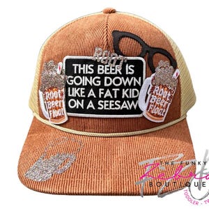 May include: A brown corduroy trucker hat with a tan mesh back. The hat features a black sign with the text "THIS BEER IS GOING DOWN LIKE A FAT KID ON A SEESAW". It has two "Root Beer Float" patches and a pair of black glasses.
