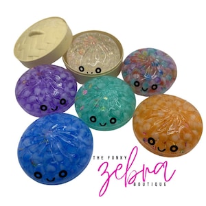 May include: A collection of colorful, round, squishy toys with smiling faces. The toys are in shades of blue, purple, green, and orange, and are filled with small, clear beads. One is in a beige box. The Funky Zebra Boutique logo is in the bottom right.