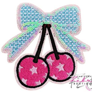 May include: A decorative patch featuring a bow and cherries. The bow is light blue with iridescent sequins, and the cherries are pink with white star accents. Black stems and a white outline complete the design.