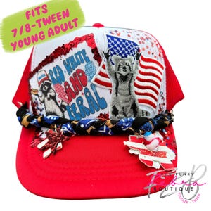 May include: Red and white trucker hat with a red brim and white mesh back. The hat features a patriotic design with a raccoon, American flag, and the text "Red, White, and Feral". The hat is decorated with stars and a braided band.