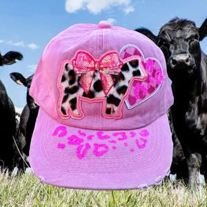 May include: Pink baseball cap with a cow design. The cap features a sequined bow and a cow-shaped patch with a black and white pattern. The brim has a pink leopard print design. Cows are in the background.