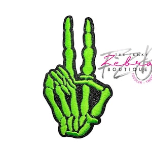 May include: A neon green embroidered patch in the shape of a skeleton hand making a peace sign. The patch has a black glitter outline and the text "THE FUNKY Zebra BOUTIQUE TODDLER TWEEN" in the upper right corner.