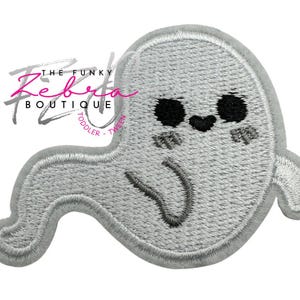 May include: Embroidered patch featuring a friendly ghost design. The ghost is light gray with black eyes, rosy cheeks, and a curved mouth. The patch has a stitched border and the text "THE FUNKY Zebra BOUTIQUE TODDLER TWEEN" in pink and gray.