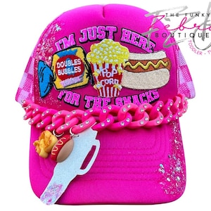 May include: A bright pink trucker hat with the text "I'M JUST HERE FOR THE SNACKS." The hat features patches of popcorn, a hot dog, and candy. A pink chain with rhinestones and food-themed charms adorns the brim. The Funky Zebeta Boutique.