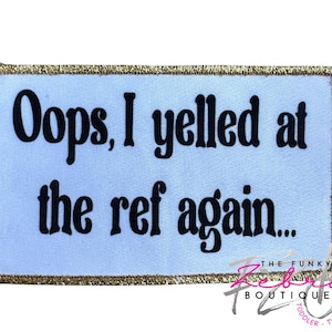 May include: Rectangular patch with the text "Oops, I yelled at the ref again..." in black font on a light blue background. The patch has a gold glitter border and a logo in the bottom right corner.