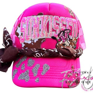 May include: A hot pink trucker hat with the word "SunKissed" in silver sequins. The hat features a brown cow print design, a brown and white bandana, and a cartoon cowgirl graphic. The Funky Tee Boutique logo is visible.