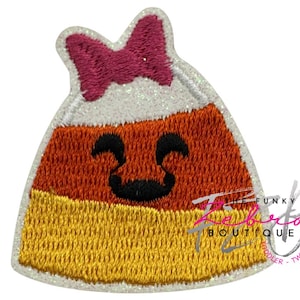 May include: Embroidered patch in the shape of a candy corn with a smiling face. The patch features a pink bow, white glitter, orange, yellow, and black colors. The text "The Funky Rebekah Boutique" is visible.