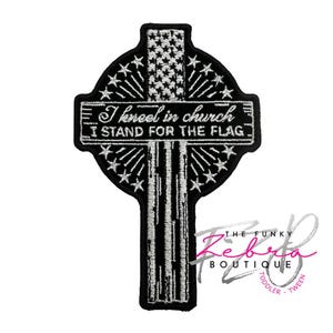 May include: Black and white embroidered patch in the shape of a Celtic cross. The top portion features a design resembling the American flag. The text "I kneel in church, I stand for the flag" is displayed. The Funky Zebra Boutique logo is in the bottom right corner.