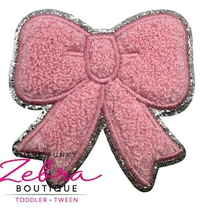 May include: A pink bow-shaped patch with a textured surface and a silver glitter border. The patch has a stitched outline and the text "THE FUNKY Zebra BOUTIQUE TODDLER • TWEEN" in pink and black.