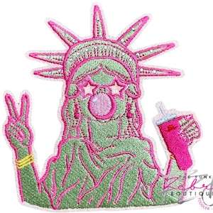 May include: Embroidered patch featuring the Statue of Liberty with a fun, modern twist. The statue is green with pink accents, wearing star-shaped sunglasses and blowing a bubble. She holds a pink drink and makes a peace sign.