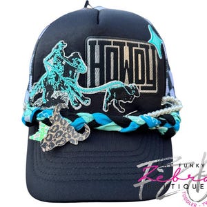 May include: Black trucker hat with a mesh back and a curved brim. The front features a turquoise and black embroidered cowboy and bull design, with the word "HOWDY" in silver. A braided band with a leopard print charm and faux pearls accents the brim.