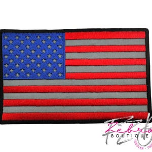 May include: Embroidered patch featuring the American flag. The flag design includes a blue field with white stars, red and gray stripes, and a black border. The patch is rectangular and suitable for application on clothing or accessories.